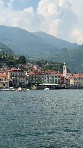 The video begins with a wide shot of a body of water, likely a lake, with a town situated on the opposite shore. The town is characterized by buildings with terracotta roofs and a prominent bell tower. Behind the town, lush green mountains rise under a partially cloudy sky. The camera then pans slowly to the right, revealing more of the town and the mountainous landscape. As the camera continues to pan, a small black motorboat with two people onboard appears in the foreground, moving across the water. The boat has a canopy and an outboard motor. The town and the mountains remain in the background throughout the panning motion.