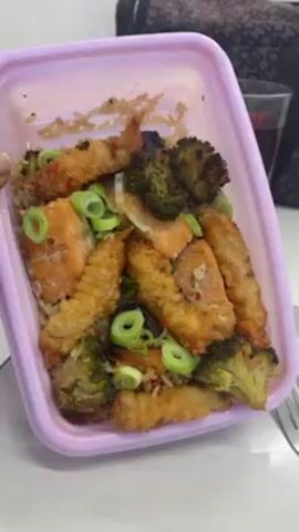 A person is shown holding a pink plastic container with a meal inside. The meal appears to consist of salmon, tempura shrimp, rice, and broccoli, garnished with sliced scallions. The person tips the container over, dumping the meal onto a white plate. There is a fork next to the plate and a glass in the background.