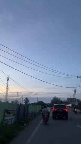 This is a vertical video shot from a moving vehicle on a road in Banda Aceh, Indonesia. The time of day appears to be dusk or early evening, indicated by the soft, diffused light and the clear blue sky transitioning to a lighter hue near the horizon. There are several utility poles and overhead wires visible, typical of urban or semi-urban areas.

In the foreground, two people are riding a motorcycle, with the rider wearing a light-colored shirt and the passenger wearing a darker shirt and a helmet. A dark-colored SUV is following closely behind the motorcycle. The road has at least two lanes.

To the left of the road, there is a green fence, partially covered by some posters or banners. Behind the fence, there is vegetation, including palm trees, and a tall cell tower. The overall atmosphere is calm and ordinary, depicting a typical commute during the evening.

In the distance, a small airplane is visible in the sky, adding a subtle detail to the scene. The weather appears clear with some faint clouds. There is no discernible text in the video that is clear enough to read. The focus of the video is primarily on the road and the sky, capturing a fleeting moment of travel.