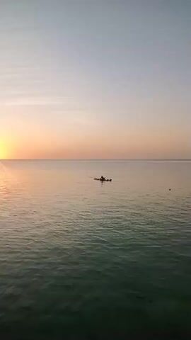 A video shows a serene ocean scene at sunrise. The sun, a bright orb, is visible on the left side of the frame, casting a golden reflection across the water. The sky transitions from a pale blue at the top to a soft orange and pink hue near the horizon. The water is calm with gentle ripples. In the middle of the frame, a lone individual is seated in a kayak, paddling across the tranquil sea. The overall atmosphere is peaceful and tranquil.