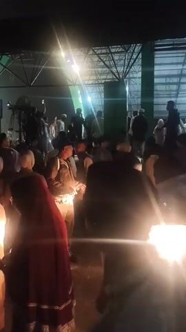 A crowd of people, some wearing traditional Islamic attire, are gathered indoors under artificial lighting. In the background, a banner with text in what appears to be Indonesian is visible, along with a large green structure. Several individuals in uniform, possibly security personnel, are present among the crowd. The scene suggests an event or gathering is taking place, possibly a religious or community event, with a stage or focal point visible in the distance. The lighting is bright and somewhat dramatic, creating lens flares and highlights.