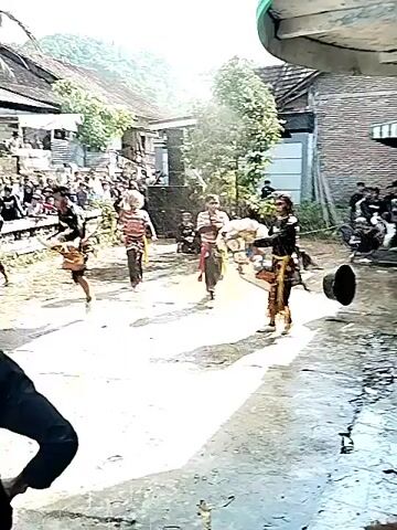 This video captures a traditional Jathilan or Kuda Lumping dance performance in a village setting in Jenawi, Indonesia. The event takes place outdoors on a sunny day, with a clear sky. The performance area is a mix of dirt and concrete, with some muddy patches.

Several performers are visible. Some are dressed in traditional costumes, including two dancers with hobby horses, one of whom is also wearing a mask resembling a barongan. Another performer, shirtless and wearing traditional lower garments, crawls on the muddy ground. A dancer in a black shirt holds and plays a black drum, while another in a similar outfit carries a white, fan-like object. The dancers move energetically to traditional music, with visible drums and possibly other gamelan instruments contributing to the sound.

A crowd of spectators, including men, women, and children, watches the performance from behind a low stone wall and from various vantage points around the open area. Many young men in the foreground are wearing matching black t-shirts with yellow text. The text on their shirts includes "RANG'90'S" and "Sakjane malah... RANG'90'S". A banner in the background is partially visible, also featuring "RANG...". Houses and lush green trees line the background, suggesting a rural or semi-rural environment, possibly in a hilly or mountainous region. The overall atmosphere appears lively and culturally rich, with the audience engaged in watching the performance.