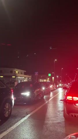 The video captures a nighttime scene of a street lined with parked and moving cars. In the foreground, the taillights of a dark-colored car are prominently visible on the right, illuminated by red light. To the left, the side of a black SUV is seen. The road is wet, reflecting the streetlights and the headlights of the vehicles. A line of cars is queuing on the road, suggesting either a traffic jam or a gathering. The cars are modern, with some appearing to be luxury models.

In the background, there are buildings with illuminated windows, possibly shops or residences. Several streetlights illuminate the scene. A few people are visible standing near the buildings on the left, appearing to be gathered. The sky is dark, indicating it is nighttime, with some faint lights or reflections visible in the upper portion of the frame. The overall atmosphere suggests an urban or suburban environment at night, possibly a social gathering or a busy street.