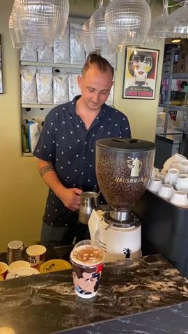 A man stands behind a counter in what appears to be a coffee shop. He is wearing a dark blue short-sleeved shirt with a white pattern of palm trees. He has a man bun and a mustache. He is holding a metal milk pitcher in his left hand. In front of him is a large white coffee grinder filled with coffee beans, with the brand name "HAUSBRANDT" visible. To the right of the grinder are stacks of white ceramic cups. On the counter in front of the man and the grinder is a clear plastic cup with a beverage inside, topped with foam and caramel drizzle. Behind the man, shelves are visible, stocked with bags of coffee. A framed poster with a graphic of a woman's face and text is hanging on the wall. There are also several clear glass items suspended from the ceiling above.