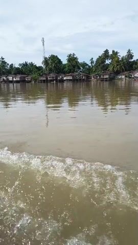 The video shows a view from a moving boat on a wide, murky river. In the foreground, white waves are crashing against the side of the boat, indicating movement. In the background, a shoreline is visible with a line of stilted houses, some with corrugated metal roofs, and lush green trees, including palm trees. A tall cell tower stands out against the cloudy sky behind the houses. The overall impression is of a rural waterside community.