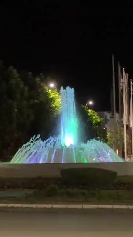 The video shows a brightly lit fountain in the center of a roundabout at night. The fountain is lit with green, blue, and white lights. Trees and streetlights surround the fountain.  Several tall flagpoles stand near the fountain. A road with cracked asphalt is visible in the foreground. Cars drive in the distance. The sky is completely dark.