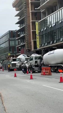 The video captures a construction scene on a street in Coquitlam, Canada, during the daytime under an overcast sky. Multiple white concrete mixer trucks are parked in a line along the side of the road, which is sectioned off by orange traffic cones and barriers.

A tall, multi-story building under construction, with visible scaffolding and unfinished floors, dominates the right side of the frame. To the left, another modern-looking building with numerous windows is visible. Several construction workers are present on the street. In the foreground, a worker wearing a black jacket with high-visibility yellow stripes and a hard hat walks towards the camera. Other workers are seen near the parked concrete trucks.

Visible text on the trucks includes "LAFARGE" on one and "AMRIZE" on the last truck in the line. The scene suggests ongoing construction activity, likely involving concrete pouring for the new building.