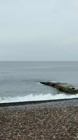The scene is a calm, overcast day at a pebble beach. The sea is a muted gray, with gentle waves lapping against the shore. A small, concrete breakwater extends into the water, partially submerged. The beach is covered with smooth, rounded pebbles in various shades of brown and gray. The sky is uniformly overcast, casting a soft, diffused light over the scene. No people are visible.