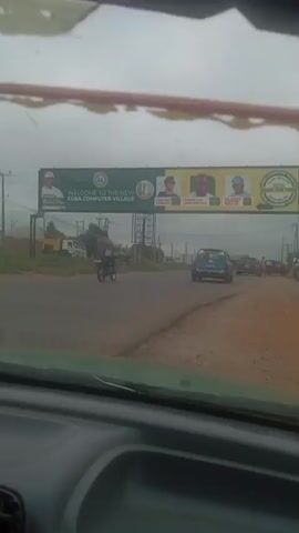 The video appears to be filmed from inside a vehicle. The perspective looks forward through the windshield, showing a road with other vehicles and a large billboard overhead. The billboard is green and yellow and reads "WELCOME TO THE NEW EGBA COMPUTER VILLAGE". It also features images of several men and a phone number. The sky is overcast. Traffic is visible on the road, including cars and motorcycles. The environment appears to be semi-urban or rural, with some vegetation and structures visible in the background.