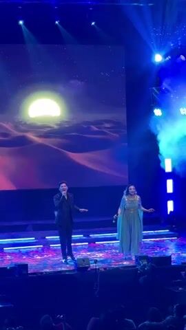 Two singers are performing on stage in front of a large audience. The male singer is wearing a black suit and holding a microphone in his right hand. The female singer is wearing a long, pale blue dress and also holding a microphone. They are standing on a stage with a colorful patterned floor, and there are blue lights shining around them. Behind them, a large screen displays an image of a desert landscape under a starry night sky with a large, bright moon. There is smoke or fog on the right side of the stage. The audience is visible in the foreground, some holding up phones or cameras.