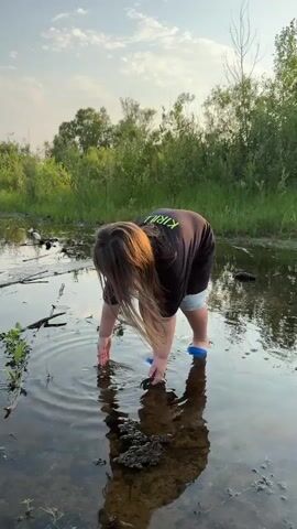 A young child with long brown hair, wearing a black t-shirt with a green alien graphic and light blue denim shorts, stands in shallow, murky water. Blue sandals are on the child's feet. The child is bending over, reaching a hand into the water. The background consists of green foliage and trees under a pale sky. There are some sticks and debris in the water. The child's reflection is visible on the surface of the water.
