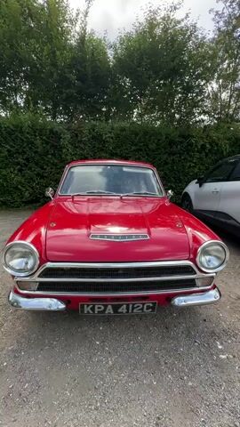 The video shows a red vintage Ford Cortina car parked outdoors. The car is viewed from the front, with a slight pan to the side. It has chrome bumpers and trim. The license plate reads KPA 412C. A white modern SUV is parked to the right of the vintage car. The background consists of a green hedge and trees. The ground is covered in gravel. The video is brief, lasting only a few seconds.