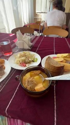 The video shows a meal being served in a brightly lit dining hall with a checkered tile floor and yellow and brown pillars. The tables are covered with maroon and white striped tablecloths. In the foreground, a table is set with a bowl of soup with croutons and potatoes, a plate of salad, a plate with bread and sliced apples, and a glass of red liquid. A fork and knife are visible on the table. In the background, several people are seated at tables, some eating and others conversing. The camera pans to show more of the dining hall and the other patrons. One woman, wearing a white shirt, is seated at a table in the middle ground, with her back to the camera. Further back, a woman with blonde hair and sunglasses is smiling and looking at her phone while seated at a table. The overall atmosphere appears casual and communal.