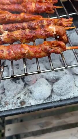 The video shows several rows of seasoned meat skewers grilling over hot coals. The skewers are arranged horizontally on a metal grill rack. The meat appears to be thinly sliced and wrapped around wooden skewers, coated in a reddish-orange spice mixture. Some of the meat is beginning to char and caramelize. To the right of the skewers, pieces of chicken, including drumsticks and thigh portions, are also grilling. The chicken pieces are visibly browned and have grill marks. The coals below are glowing and covered in a layer of white ash. The overall scene depicts an outdoor barbecue in progress.