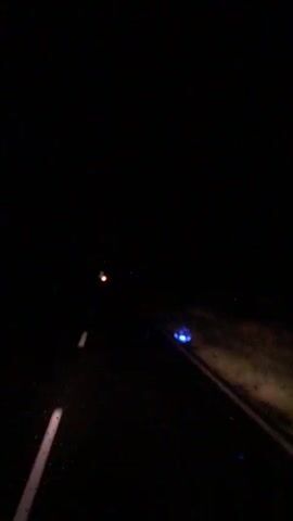The video shows a first-person perspective of driving at night. The road ahead is dark, with white lane markings visible. There are a few lights in the distance, including a red light and a blue light on the right side of the road. The environment appears to be rural or undeveloped, with sparse vegetation visible on the sides of the road. The lighting is poor, suggesting a lack of streetlights or other artificial illumination. The camera is shaking slightly, indicating movement, likely from the vehicle. There are no people or other vehicles clearly visible in the frame.