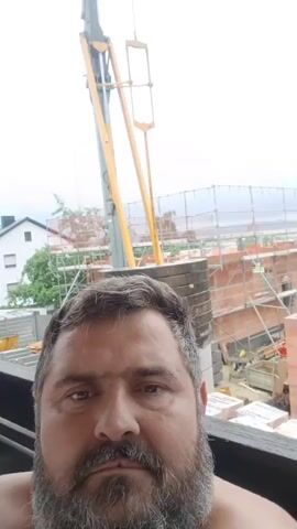 A man with a beard and wide eyes makes a surprised face into the camera, held at a slightly low angle. Behind him, a construction site is visible with scaffolding and stacks of bricks. A yellow crane arm extends upwards into the sky, with a mirrored object at its tip. The sky is overcast.