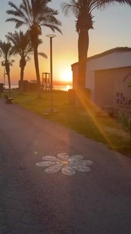 The video opens with a shot of a palm tree silhouetted against a sunrise. The camera pans to the right, revealing a building with a sign that reads "Hurma, Cafe, Bar, Restaurant." A Buddha statue is visible in the foreground, along with a wooden chair and lush greenery. The camera then moves to a large mirror reflecting a woman standing in front of it. The woman is wearing a black tank top, black leggings, and white sneakers, with a white jacket tied around her waist. She is holding a phone and posing for a selfie. Behind her, in the reflection, are buildings, a playground, and a fence. The video ends with a close-up of the mirror, showing the woman's reflection and the surrounding environment.