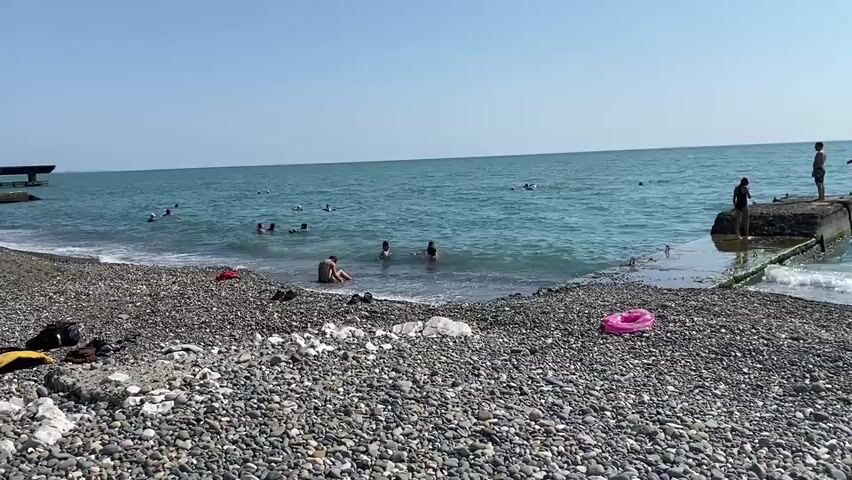 The video depicts a pebble beach with several people swimming in the clear blue sea. The water is calm with small waves breaking on the shore. In the foreground, there are various objects scattered on the beach, including bags, shoes, and a pink inflatable ring. A concrete pier extends into the water on the right side of the frame, with two individuals standing on it. Another concrete structure, possibly a breakwater, is visible in the distance on the left. The sky is clear and blue. The overall scene suggests a peaceful and sunny day at the beach.
