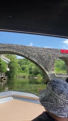 This video shows a boat ride on a body of water, likely a river, with a large, old stone bridge in the background. The bridge has two arches and appears to be made of rough-hewn stones. A red flag with a golden emblem, likely a national flag, is displayed on the bridge. 

Two individuals are visible in the foreground, seen from behind, wearing bucket hats. The person on the left wears a white bucket hat with a black paisley pattern. The person on the right wears a dark grey bucket hat. They are seated on a boat, with a dark canopy overhead providing shade. The water is calm and reflects the sky and surrounding greenery. 

The scene suggests a sunny day with clear blue skies and fluffy white clouds. Lush green trees and vegetation line the banks of the waterway, creating a picturesque natural setting. The overall atmosphere appears relaxed and peaceful, typical of a scenic boat tour. The location is identified as Rijeka Crnojevića, Montenegro. There is no visible text in the video.