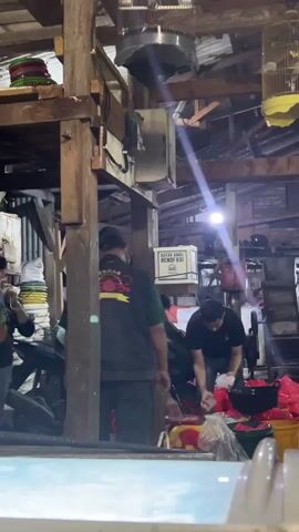 This is a vertical video clip showing an indoor scene with several people. The setting appears to be a workshop or storage area, with wooden beams and corrugated metal walls. In the foreground, a blurry light blue surface is visible, possibly a counter or table.

The main action focuses on several men. On the right side of the frame, two men are actively engaged with red and yellow plastic bags, and what appears to be a white bucket containing a white substance, possibly ice. One man is wearing a black t-shirt and is reaching into the bucket, while the other, in a grey shirt, is also near the bags.

To the left, a man in a green t-shirt is standing and looking towards the center. Another person, wearing a black shirt and grey pants, is bending down near a stack of items. A man in a red shirt is partially visible behind a wooden pillar. In the background, there are several white boxes, one of which has text that is partially legible but appears to be Indonesian. Bird cages are suspended from the ceiling. The lighting suggests it is daytime, with a bright light source creating a visible beam.