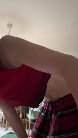 A person wearing a red top and plaid shorts is bending over and touching their toes. The person has a watch on their left wrist. In the background, there is a table and chairs, as well as a hanging light fixture on the ceiling. The scene is indoors.