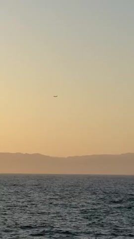 A vast expanse of ocean stretches across the bottom of the frame, its surface gently rippling under an overcast sky. In the distance, a hazy silhouette of mountains runs along the horizon, their peaks softened by atmospheric perspective. Above the mountains, the sky transitions from a pale yellow near the horizon to a lighter hue towards the top of the frame. High in the sky, a small airplane is visible as a dark silhouette, its wings extended as it traverses the frame from left to right. The overall scene is serene and tranquil, evoking a sense of open space and quiet movement.