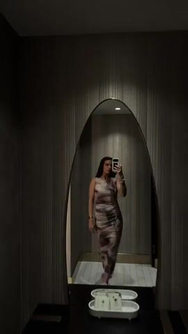 A woman stands in front of a large oval mirror, taking a selfie with her phone. She is wearing a long, form-fitting dress with a tie-dye pattern. The dress is sleeveless and appears to be made of a silky material. The woman has long, dark hair that falls around her shoulders. She is wearing bracelets on both wrists and rings on her fingers. Her nails are painted dark. Her makeup appears to be minimal. The mirror is framed by a dark, curved edge, and reflects the woman and her surroundings. The room appears to be a hallway or dressing room, with textured walls and a checkered floor. There are lights visible on the ceiling. The woman poses in various ways, looking at herself in the mirror and holding her phone up to take the photo.