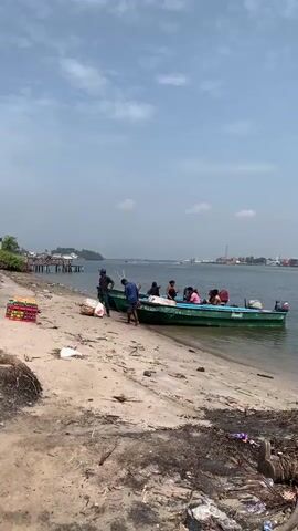 A small, green motorboat with an outboard engine is partially beached on a sandy and muddy bank alongside a wide body of water. Several individuals, approximately 6-8 people, are gathered around and in the boat. Initially, two men stand near the bow, one in a black shirt and another in a blue shirt, while others are seated inside the vessel.

The water is calm, reflecting the partly cloudy sky. The bank is visibly littered with debris and refuse. In the background, across the water, a distant shoreline reveals an industrial complex. Prominent features include several large, white cylindrical storage tanks, various buildings, and what appear to be cranes, indicating a port or industrial area.

As the video progresses, the boat pushes off from the bank and moves further into the water. All the individuals are now seated within the boat as it begins to travel across the waterway. The scene depicts a routine activity of water transport or movement of people in a bustling riverine or estuarial environment. The time of day is clearly daytime, with bright ambient light and good visibility. No discernible text is visible on the boat or in the distant background structures. The overall atmosphere suggests an everyday scene in an active waterway, possibly in the Ibasa area of Nigeria as per the provided context.