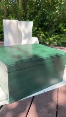 A close-up shot focuses on a dark green wooden box outdoors. The box is on a wooden table with some white paper towels beneath it. In the background, there are trees and green foliage. A person's hands appear, opening the box. As the lid is lifted, a cloud of white smoke or vapor is released from inside the box, obscuring the interior. The smoke dissipates as the lid is further opened, revealing an orange-colored interior with text that appears to be a logo and some writing. The person's fingers are visible near the edge of the lid as the smoke drifts.