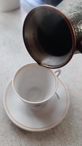 A close-up shot shows dark liquid being poured from a metal pot into a white teacup with a gold rim. The liquid appears to be hot, as steam is visible rising from the cup. The cup is sitting on a matching saucer. The background is blurred but appears to be a light-colored surface.