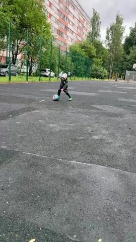 A person wearing a grey jacket and green boots is playing soccer on a paved outdoor court. The person is kicking a colorful soccer ball. The court is surrounded by a green fence. In the background, there is a red brick building, trees, and parked cars. The ground is wet, suggesting it may have recently rained.
