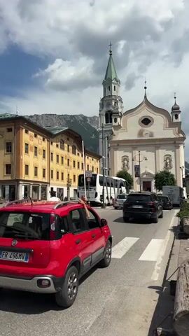 The video shows a street scene in what appears to be a European town. In the foreground, a red car is stopped at a crosswalk. Behind it, a white bus is also stopped. Further down the street, other cars and a truck are visible. To the left of the street is a yellow building with arched windows. To the right of the street is a church with a tall spire. In the background, mountains are visible. A man wearing a white t-shirt and shorts is walking across the street. Another man wearing a white hat and shorts is standing on the sidewalk. The sky is cloudy.