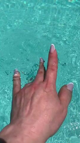 A person's hand is submerged in clear, turquoise water, creating ripples and splashes. The hand has manicured nails with a white French tip. The lighting suggests it is daytime. The water appears to be a swimming pool.