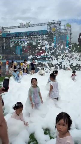A foam party is taking place outdoors in front of a stage. Children of various ages are playing in a large mound of white foam. Some children are sitting, others are standing and splashing. In the background, a stage structure with the words "WINDOW OF THE WORLD" is visible, along with scaffolding and buildings. The sky is overcast.