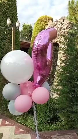 A bouquet of balloons, featuring a large pink number "9" foil balloon and several smaller latex balloons in white, gray, and pink, is displayed outdoors. The balloons are arranged in a cluster, tied together with white ribbons. They are positioned on a paved walkway with a patterned brick design.

In the background, lush green cypress trees and other foliage create a natural setting. A stone wall or rock formation is visible behind the balloons, also covered in greenery. Two outdoor lanterns with glass shades are mounted on poles, suggesting an outdoor living space or garden area. The sky is partly cloudy, indicating it is daytime, likely late afternoon due to the soft lighting. The overall impression is festive and celebratory, possibly for a birthday or special occasion. There are no people visible in the frame. The location is identified as Buzovna, Azerbaijan.