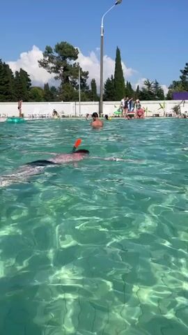 A person wearing a snorkel mask and orange mouthpiece swims in a bright turquoise swimming pool on a sunny day. They are on the left side of the frame, facing away from the camera and towards the right. The water is clear, allowing the tiled bottom of the pool to be seen. In the background, several other people are visible in and around the pool, some sitting on lounge chairs and others standing. There are trees and a white fence bordering the pool, with a clear blue sky above. The overall scene depicts a recreational setting at a pool.