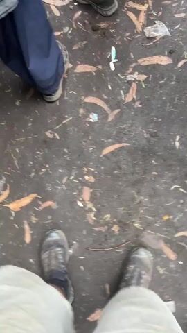 The video shows a view of a person's feet wearing boots standing on a wet, muddy ground. The ground is littered with dried leaves, twigs, and a few pieces of trash, including what appears to be a plastic wrapper and a white object that may be a used toothbrush. In the background, other people's legs and feet are visible, wearing various types of footwear including sneakers and boots. One person is wearing dark blue pants and a grey and white striped shirt. The lighting suggests an outdoor environment, possibly overcast or in shadow. The perspective is a first-person view, looking down at the ground.