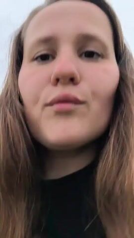 The video begins with a close-up of a woman's face, looking directly at the camera. She has long brown hair and is wearing a dark top. The background is a blurred landscape of green fields and a cloudy sky. As the video progresses, the camera pans out slightly, revealing more of the environment. The woman is standing in a grassy field with a dirt path winding through it. In the distance, there are trees and rolling hills. The woman occasionally smiles and gestures towards the scenery, seemingly sharing her surroundings with the viewer. The overall atmosphere of the video is casual and personal, suggesting a vlogging or personal update style.