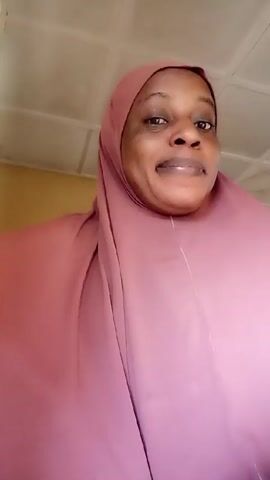 A woman wearing a pink hijab smiles at the camera. She is indoors. The ceiling is visible. The wall is yellow.