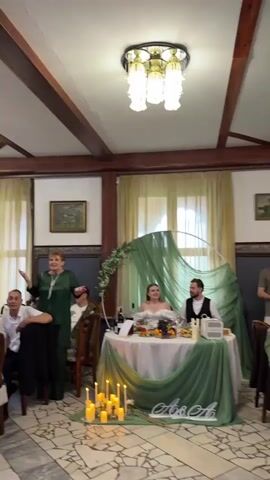 A group of people are gathered in what appears to be a banquet hall. A bride and groom are seated at a decorated head table in the center of the room. A woman in a green outfit stands and addresses the room. Several other guests are seated at tables and some are standing. The room has wooden beams on the ceiling and a chandelier. There is a semi-circular arch decorated with fabric and greenery behind the bride and groom. Candles are lit on the tables. A man in a suit is holding a microphone and speaking to the crowd.