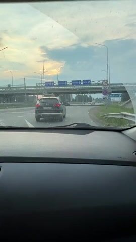 The video shows a view from the inside of a car looking through the windshield. The car is driving on a road. In front of the car, there is another black SUV and a white van. Ahead, there is an overpass with signs that read "PERM" and "OSA". The sky is cloudy with some sunlight visible. There are streetlights and traffic signs on the side of the road. The road has multiple lanes, and the car appears to be in one of the right lanes. The dashboard of the car is visible in the foreground.
