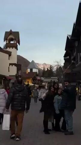 The video shows a pedestrian street in what appears to be a European-style village or resort town. The street is paved with cobblestones and lined with buildings, some with traditional Alpine architecture, featuring gabled roofs and wooden details. A tall, light-colored tower with a bell or viewing platform stands on the left side of the street. In the background, snow-capped mountains are visible under a light-colored sky. Several people are walking along the street, and a small group of four people is taking a selfie with a smartphone. One man is holding the phone, and the other three are posing with him. Some people are carrying shopping bags. The overall atmosphere seems relaxed and touristy.