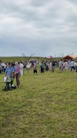 The video shows an outdoor gathering with several tents and a large grassy field. People are milling around, some standing in groups, others walking. In the foreground, three people are walking towards the camera: a young girl in a white t-shirt and shorts, a woman in a tan suit and white top, and an older woman in a plaid shirt and jeans. Further in the background, a person in a wheelchair is being pushed by another individual. Several tents are visible, suggesting a market, festival, or fair. The sky is cloudy, and the overall atmosphere appears to be casual and public.