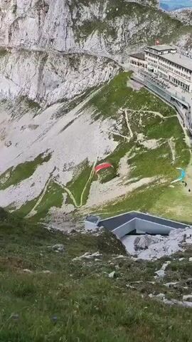The video captures an aerial view of a mountainous landscape with paragliders. In the foreground, lush green grass covers a steep slope. Further up the mountain, a paraglider with a red canopy is visible, soaring against a backdrop of rocky terrain and sparse vegetation. A winding path is discernible on the mountainside. In the distance, a large building, possibly a hotel or a research facility, is perched on the mountain. Another paraglider, with a blue and white canopy, is seen further away. The sky appears clear, suggesting good weather conditions for paragliding. The overall scene depicts a recreational activity in a dramatic natural setting.