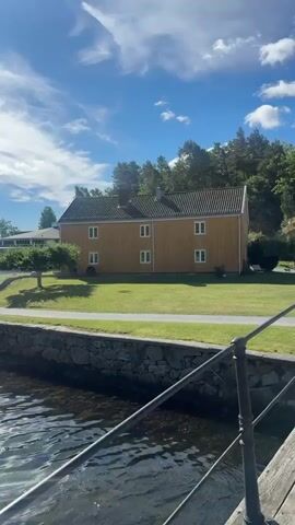 The video opens on a bright, sunny day with a blue sky dotted with white clouds. In the foreground, the choppy, sun-dappled surface of a body of water is visible, with a stone embankment to the right. Beyond the embankment, a neatly manicured green lawn stretches towards a traditional two-story yellow house with white-trimmed windows. A paved pathway runs along the edge of the lawn, and a lone figure, wearing a white shirt and dark shorts, walks along it. Further in the background, a white boat is docked, and beyond that, a hilly coastline with scattered buildings and trees lines the horizon. The scene appears peaceful and idyllic, possibly a waterfront residential area or park.