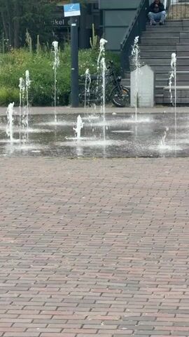 The video shows a public space with a water fountain. There are several streams of water shooting up from the ground. The ground is paved with red bricks. In the background, there are stairs leading up to a building. A person is sitting on the stairs. There is also a bicycle parked near the stairs. Green bushes and trees are visible behind the fountain. The weather appears to be overcast.