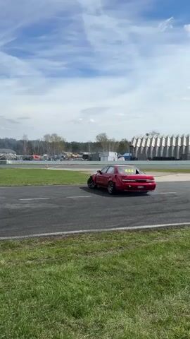 The video shows a red sedan drifting on a track. The car is missing its front bumper and has visible damage to the front end. It is labeled with the number 444. It drifts around a corner of the track, kicking up smoke from the tires. The track is surrounded by grass and has various buildings and structures in the background under a partly cloudy sky.