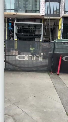 The video captures a daytime view of an active construction site, filmed from behind a black mesh fence. In the background, a multi-story building is under construction, featuring exposed metal frameworks, large window openings, and partially installed cladding. At the beginning of the video (0:00-0:01), a worker in blue overalls is visible on a blue scissor lift positioned on the left side of the building. Metal chains hang from an upper level of the structure.

The foreground is defined by a concrete sidewalk and a black mesh construction fence. The fence prominently displays the white logo "onni group" alongside a registered trademark symbol (R). From 0:01 to 0:04, a construction worker is seen behind this fence. The worker is wearing a yellow-green high-visibility jacket and a hard hat, looking upwards, and appears to be operating a piece of equipment or a control panel with their hands. A red traffic cone is visible near the right side of the fence, and orange markings are on the concrete ground. The scene depicts ongoing construction activities.
