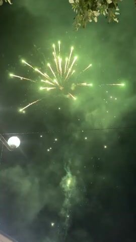 The video shows a nighttime scene with fireworks illuminating the sky. In the foreground, there are silhouetted tree branches at the top. A lamppost with a white globe is visible on the left side of the frame. The sky is dark, with bursts of light from the fireworks in various colors, including green, gold, and red. There are also hints of pink and purple smoke or haze low in the frame, suggesting the fireworks are close to the ground or reflecting off something. The fireworks are exploding in a radial pattern, creating streaks of light against the darkness.