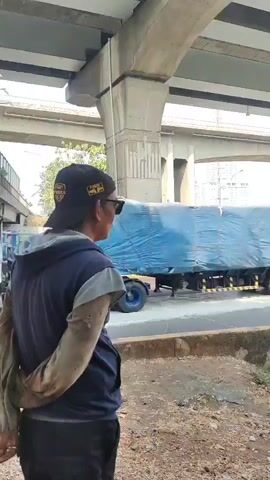 A medium shot video captures a man standing on the side of a road, looking towards traffic passing under an elevated highway structure. The man is wearing a dark baseball cap, sunglasses, and a blue hooded jacket over a dark garment. He has his back to the camera, with his hands clasped behind him. To his right, a blue-wrapped truck trailer with multiple axles and wheels is visible. The road is asphalt with yellow and white lane markings. Beyond the road, there is an embankment with overgrown vegetation and some scattered trash. The elevated highway consists of large concrete pillars and multiple levels of overpasses. In the background, tall buildings and trees are visible under a clear sky. The video also shows other trucks, including a white tanker truck and a green truck with a tarp-covered load, passing by on the road. A person wearing a headscarf and carrying a backpack is also briefly visible on the right side of the frame.